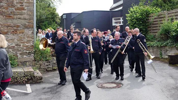 Eine Gruppe von Menschen, viele in Uniformen, marschiert mit Musikinstrumenten durch eine Straße. Zuschauer stehen am Straßenrand.