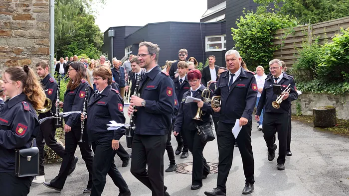 Eine Blaskapelle in dunklen Uniformen marschiert während einer Prozession auf einer Straße. Zuschauer stehen und sitzen am Straßenrand.