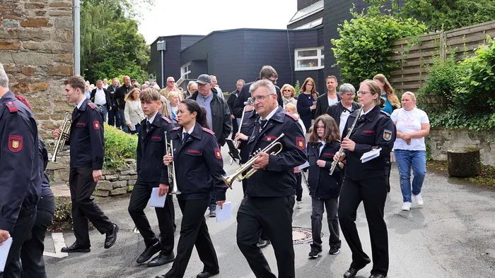 Eine Blaskapelle in dunklen Uniformen führt eine Prozession an, gefolgt von einer Gruppe von Menschen unterschiedlichen Alters und Geschlechts.