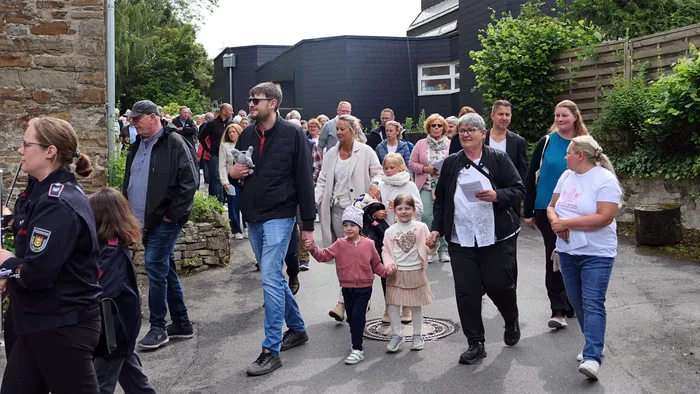 Eine Prozession mit vielen Menschen, darunter Familien mit Kindern, zieht an einem Gebäude vorbei. Eine Polizistin begleitet die Gruppe.