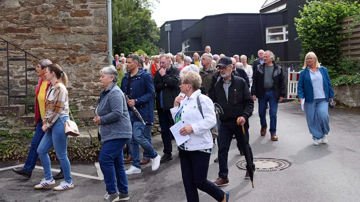 Eine große Gruppe von Menschen geht auf einer Straße, einige halten Blätter oder andere Gegenstände in den Händen. Im Hintergrund sind Gebäude und Bäume zu sehen.