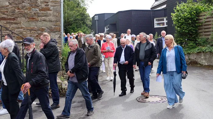 Eine Gruppe von Menschen unterschiedlichen Alters geht auf einer Straße in einer Prozession. Im Hintergrund sind Gebäude und Bäume zu sehen.