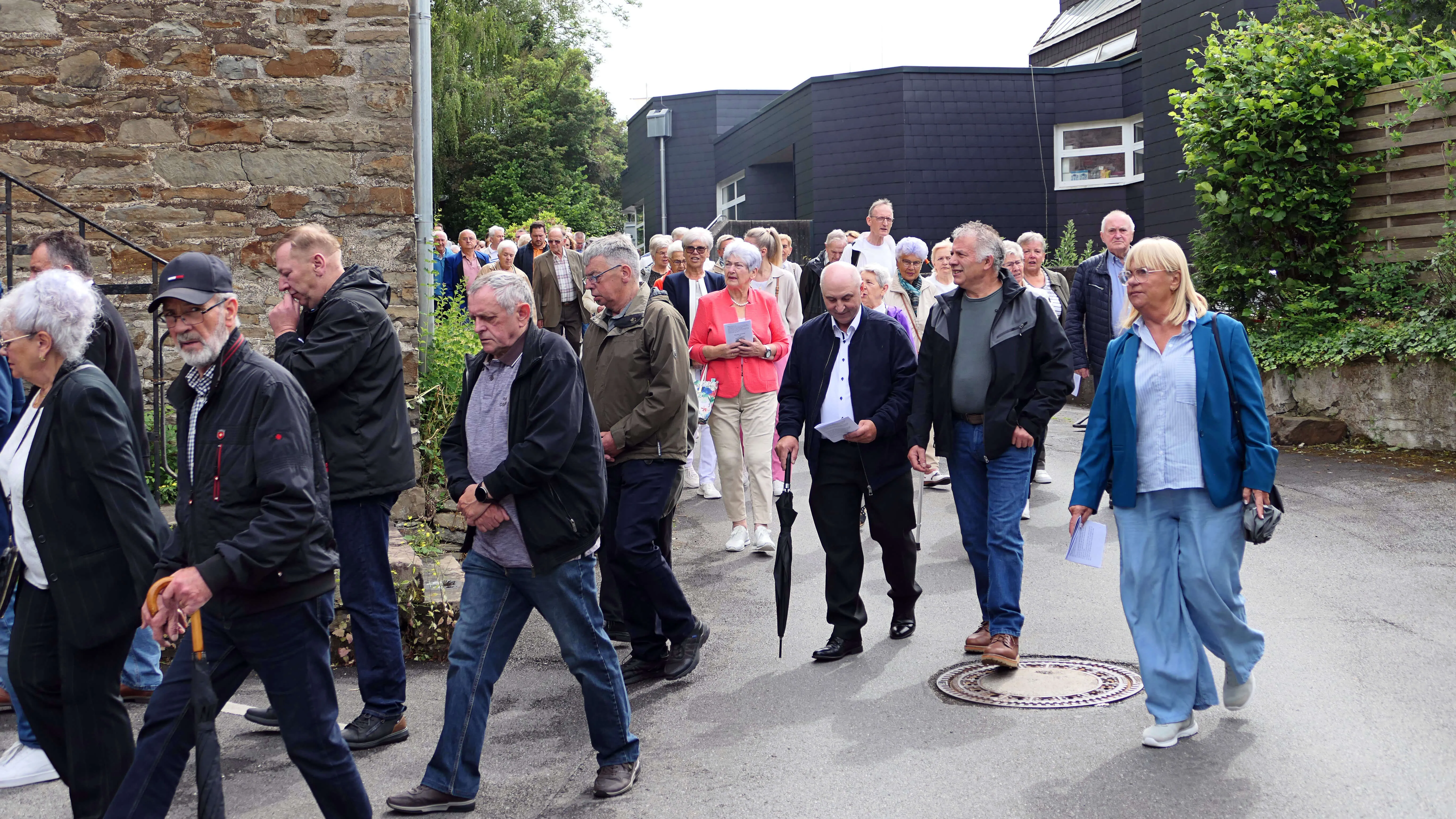 Eine Gruppe von Menschen unterschiedlichen Alters geht auf einer Straße in einer Prozession. Im Hintergrund sind Gebäude und Bäume zu sehen.