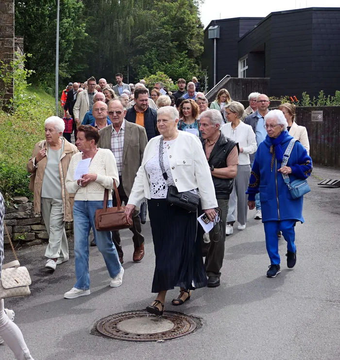 Eine Gruppe von Menschen unterschiedlichen Alters geht auf einer Straße, einige tragen religiöse Gegenstände. Im Hintergrund ein Wald und Gebäude.
