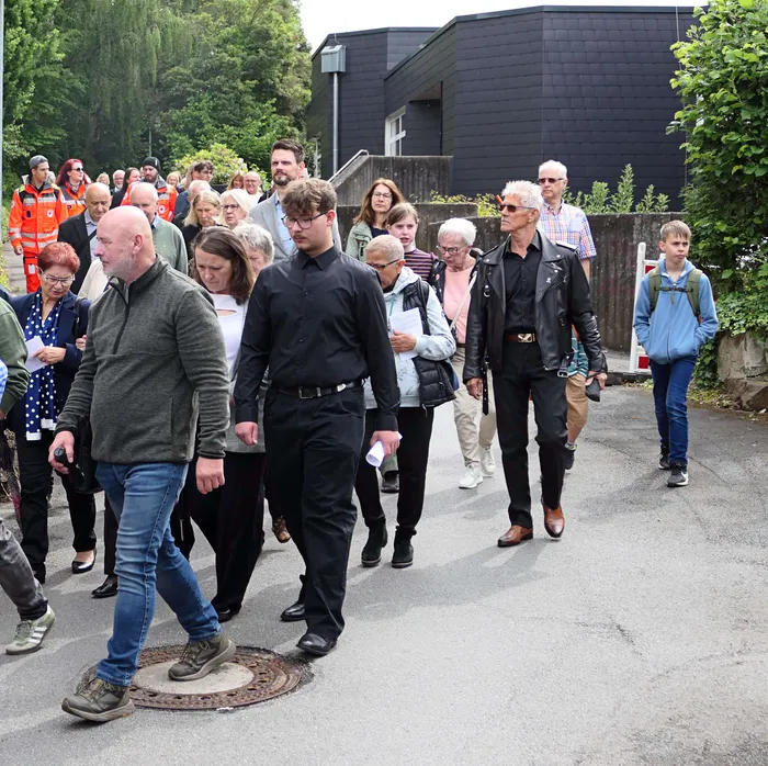 Eine Gruppe von Menschen geht auf einer Straße in einer Prozession, einige tragen religiöse Banner. Im Hintergrund ein dunkles Gebäude und Bäume.