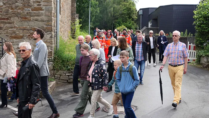 Eine Gruppe von Menschen jeden Alters geht auf einer Straße in einer Prozession, begleitet von Helfern in Warnwesten. Im Hintergrund sind Gebäude und Bäume zu sehen.