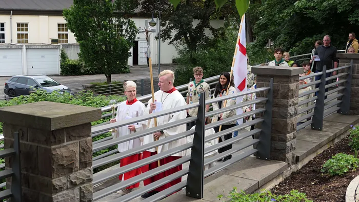 Zwei Geistliche in weißen Gewändern und rote Stolas führen eine Prozession mit Jugendlichen und Erwachsenen an, die eine Fahne tragen. Im Hintergrund ein Gebäude und Zuschauer.