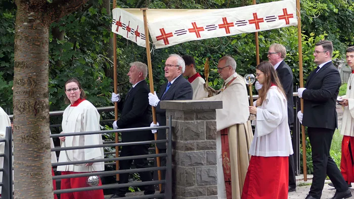 Eine Prozession mit Geistlichen und Ministranten zieht an einer Steinmauer vorbei, vornewein Banner mit roten Kreuzen auf weißem Grund.