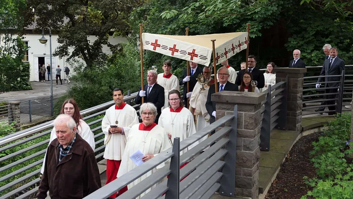 Eine Prozession mit Geistlichen und Gemeindemitgliedern schreitet vor einer Kirche entlang. Vorneweg tragen sie ein Banner und eine Monstranz.