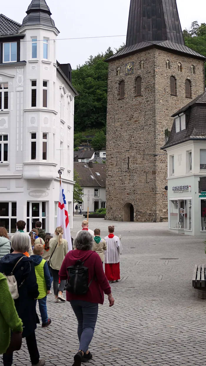 Menschen ziehen an einem mittelalterlichen Kirchturm vorbei, einige tragen Fahnen und religiöse Gewänder. Im Hintergrund sind weitere Gebäude und Bäume zu sehen.