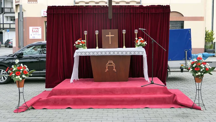 Auf einem roten Teppich steht ein Altar mit einem Kruzifix, Kerzenleuchtern und Blumen. Im Hintergrund ein roter Vorhang und ein blaues Zelt.