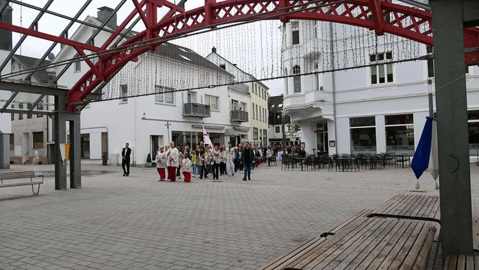 Eine Prozession mit Geistlichen und Ministranten schreitet über einen Platz vor weißen Häusern mit Fachwerkelementen. Im Hintergrund sind weitere Menschen und Cafés zu sehen.