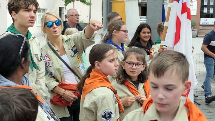 Eine Frau in Scout-Uniform weist während einer Prozession auf etwas vor sich zeigend. Um sie herum sind Kinder und Jugendliche in Scout-Uniformen zu sehen, im Hintergrund eine Menschenmenge und eine Flagge.