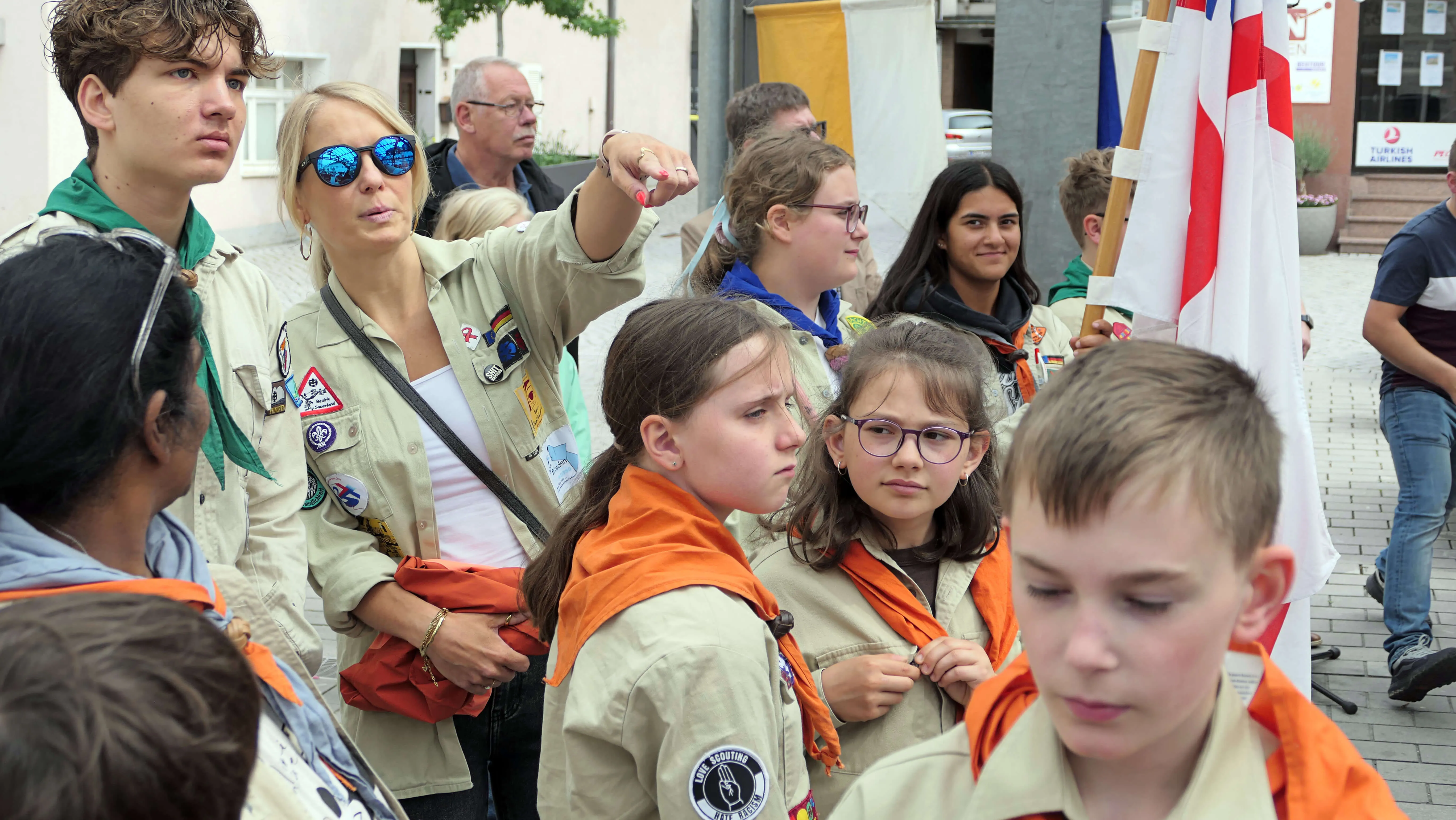 Eine Frau in Scout-Uniform weist während einer Prozession auf etwas vor sich zeigend. Um sie herum sind Kinder und Jugendliche in Scout-Uniformen zu sehen, im Hintergrund eine Menschenmenge und eine Flagge.