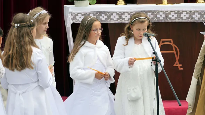 Drei Mädchen in weißen Kleidern stehen vor einem Altar und halten goldene Bänder. Im Hintergrund weitere Kinder und Erwachsene.