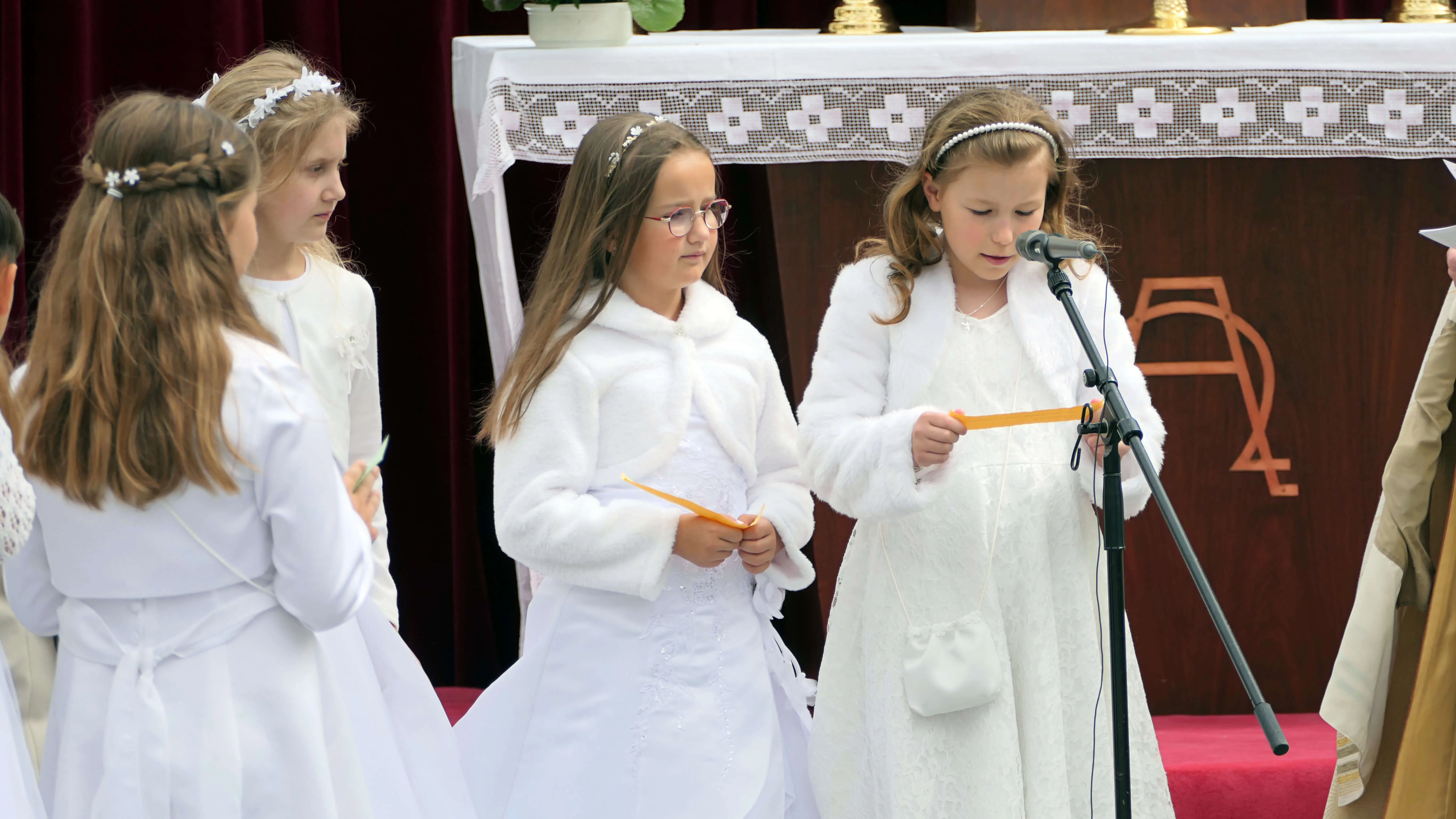 Drei Mädchen in weißen Kleidern stehen vor einem Altar und halten goldene Bänder. Im Hintergrund weitere Kinder und Erwachsene.