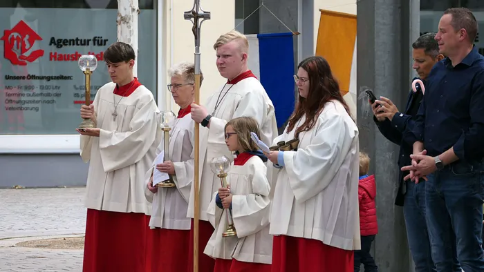 Fünf junge Menschen in roten und weißen Gewändern tragen während einer Prozession religiöse Gegenstände wie Kreuze und eine Monstranz. Im Hintergrund stehen weitere Personen und ein Gebäude mit dem Schild „Agentur für Haushalts…“.