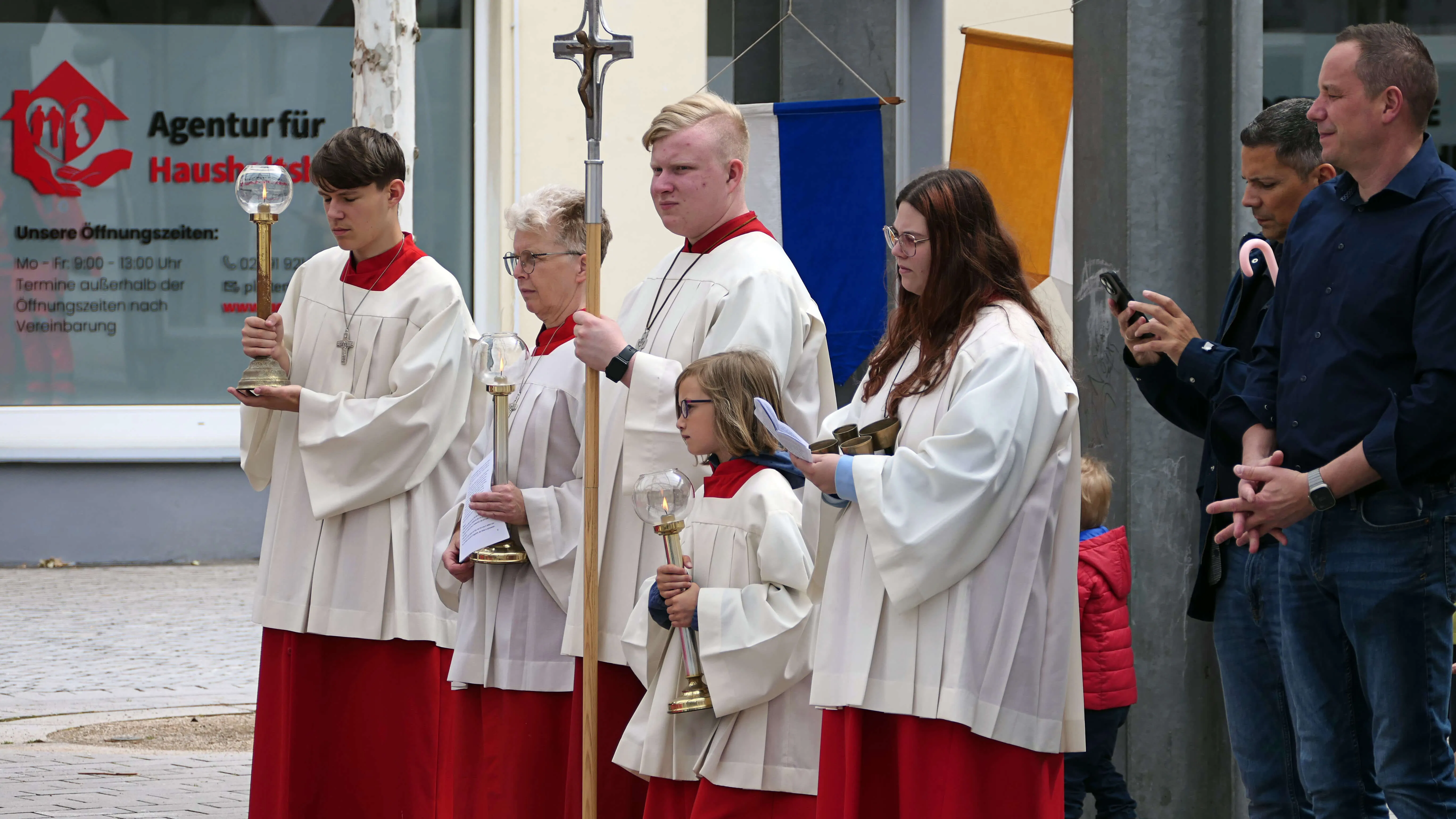 Fünf junge Menschen in roten und weißen Gewändern tragen während einer Prozession religiöse Gegenstände wie Kreuze und eine Monstranz. Im Hintergrund stehen weitere Personen und ein Gebäude mit dem Schild „Agentur für Haushalts…“.