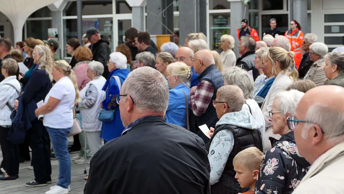 Eine große Menschenmenge steht und geht vor einem Gebäude zusammen, vermutlich während einer Prozession oder eines Festes. Im Hintergrund sind Personen in Warnwesten zu sehen.
