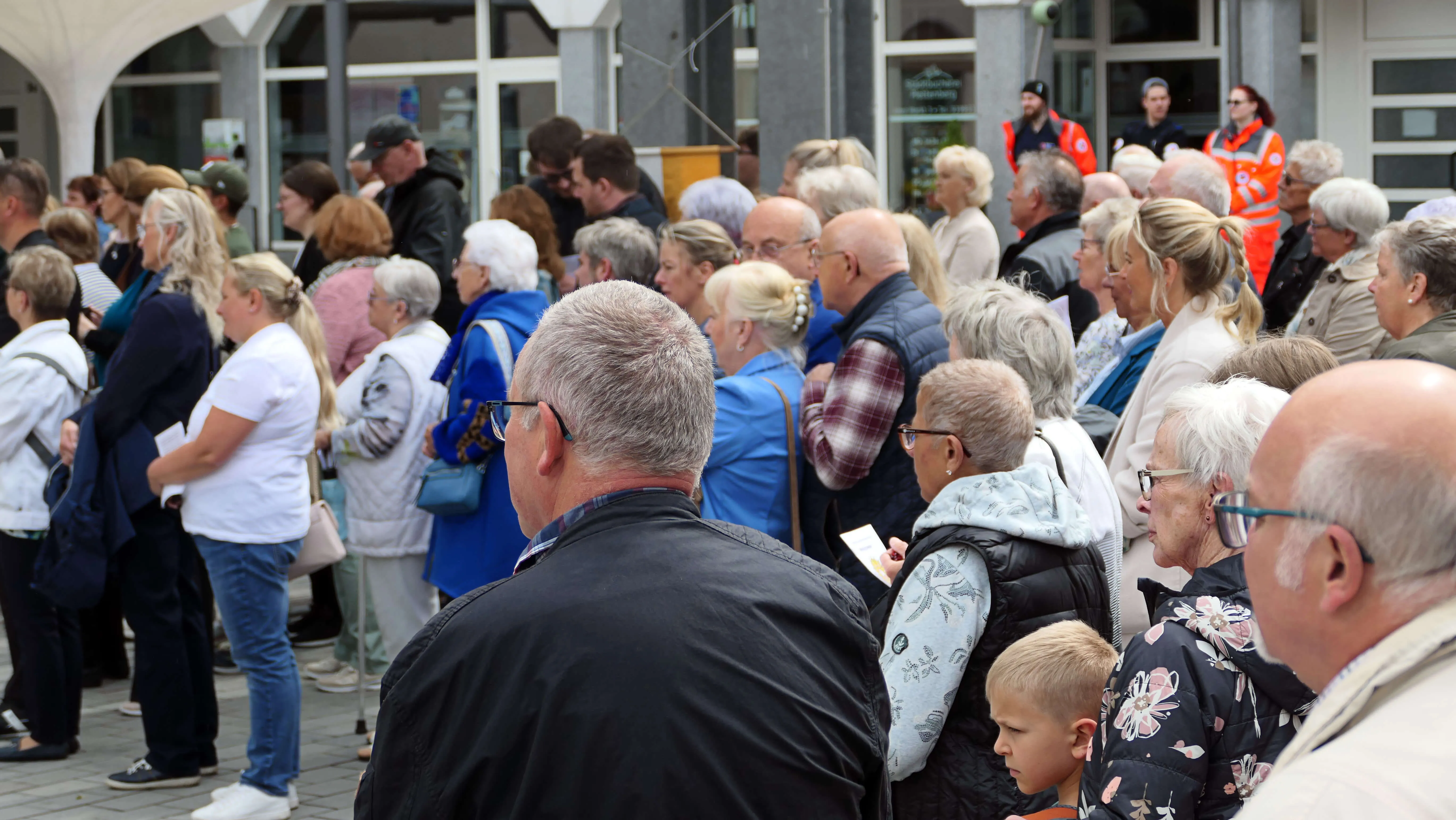 Eine große Menschenmenge steht und geht vor einem Gebäude zusammen, vermutlich während einer Prozession oder eines Festes. Im Hintergrund sind Personen in Warnwesten zu sehen.