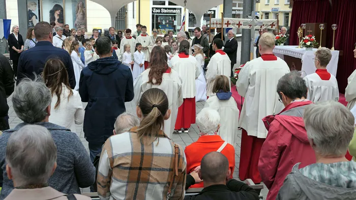 Eine Gruppe von Kindern in weißen Gewändern steht im Kreis, umgeben von Priestern in roten und weißen Gewändern, während eine Menschenmenge zusieht. Im Hintergrund sind Geschäfte und ein Banner mit der Aufschrift „Zoo Steinmann“ zu sehen.