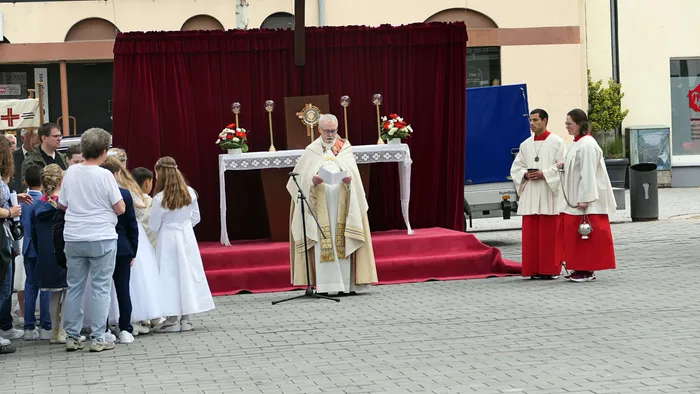 Ein Priester in goldener und weißer Gewandung hält eine Predigt vor einem Altar im Freien, umgeben von Kindern in weißen Gewändern und weiteren Gläubigen.