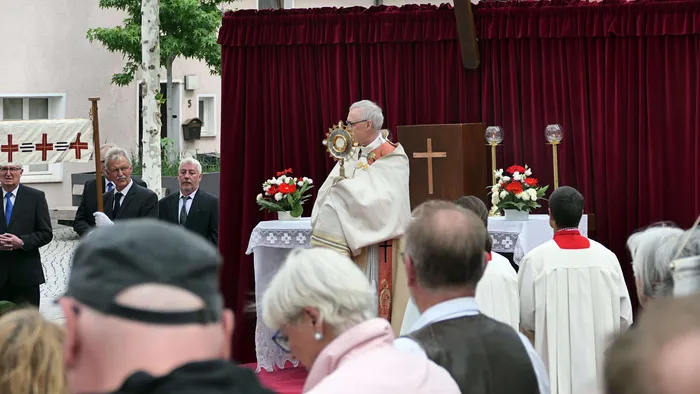 Ein Priester hält während einer Prozession die Monstranz mit der Eucharistie hoch. Im Hintergrund sind weitere Geistliche und Gläubige zu sehen.