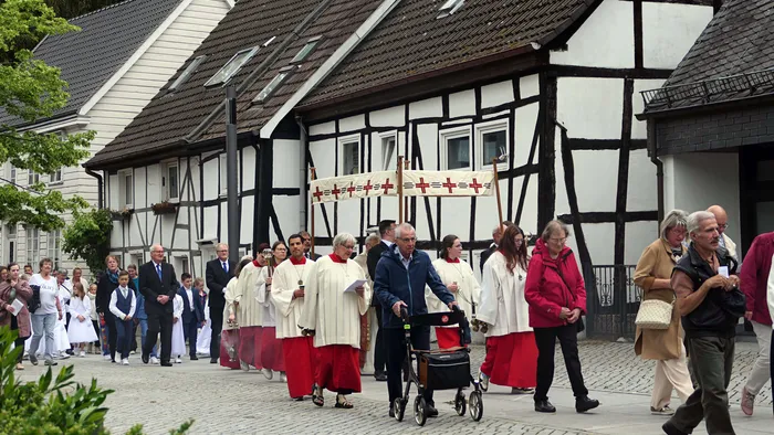 Eine Prozession mit Geistlichen in roten und weißen Gewändern zieht an einer Gruppe von Gläubigen vorbei, vor dem Hintergrund eines Fachwerkhaues.