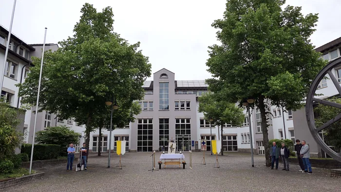 Innenhof einer Kirche mit Prozession. Im Zentrum steht ein Altar, umgeben von Fahnen und Menschengruppen.