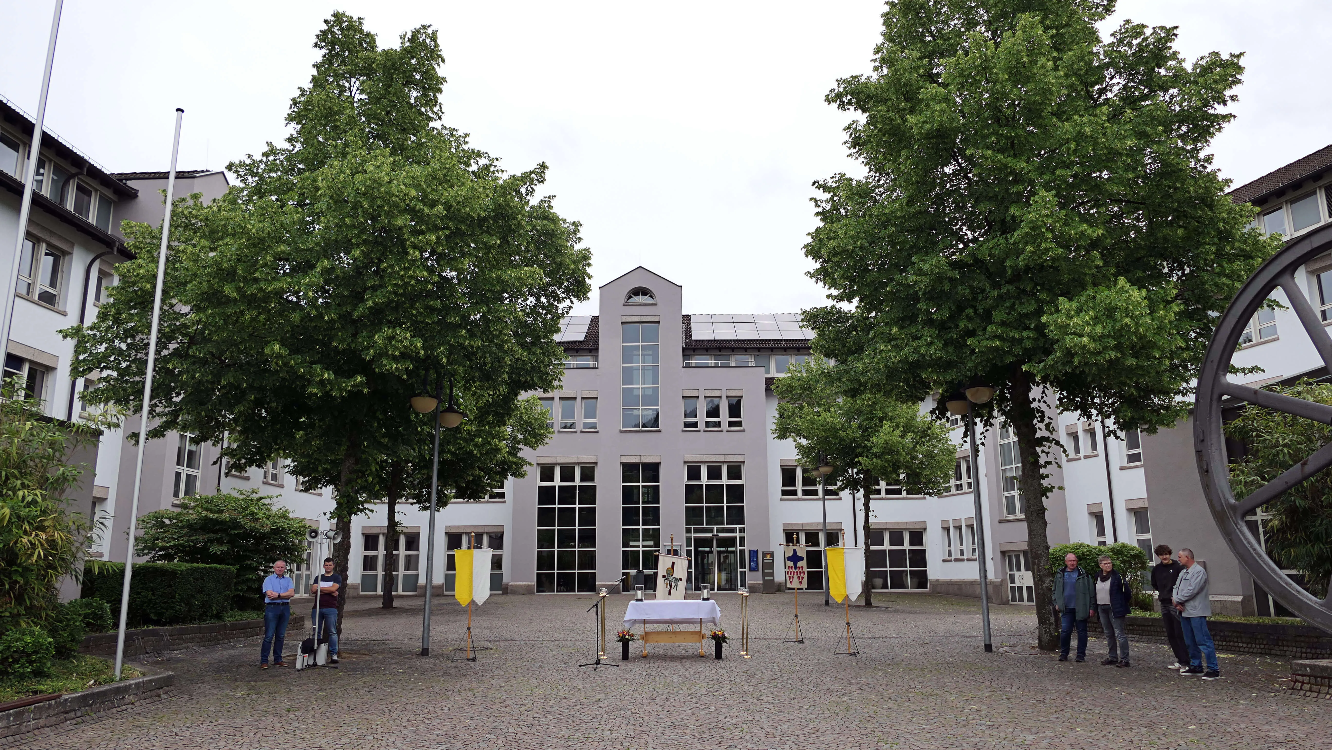 Innenhof einer Kirche mit Prozession. Im Zentrum steht ein Altar, umgeben von Fahnen und Menschengruppen.