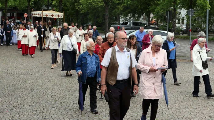 Eine Gruppe älterer Menschen zieht bei einer Prozession durch eine Stadt. Einige tragen Fahnen und halten Gebetsbücher, während sie auf einem gepflasterten Platz gehen.