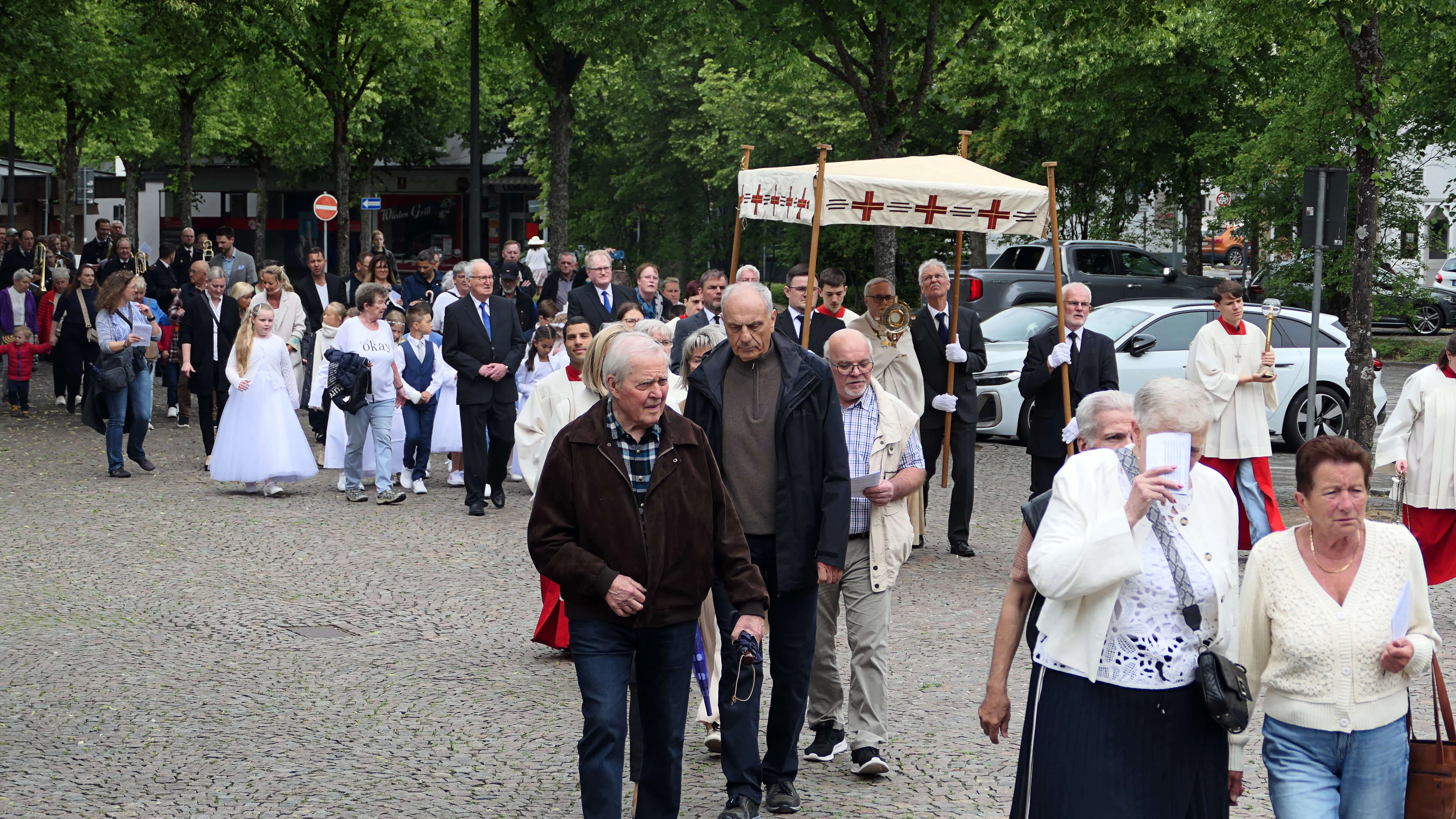 Eine Prozession mit Fahnen und Kommunionskindern zieht durch eine Stadtstraße, umgeben von Zuschauern.