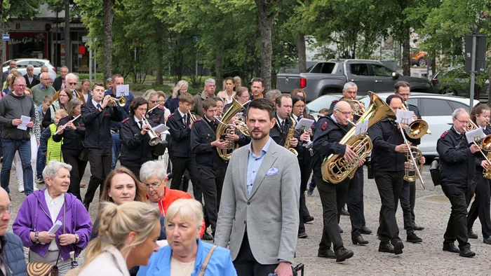 Eine Blaskapelle führt eine Prozession durch eine Stadt, begleitet von Gemeindemitgliedern. Ein Mann in einem hellgrauen Anzug geht vor der Kapelle her.