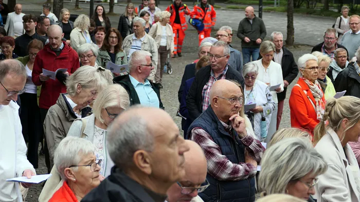 Eine große Menschenmenge steht und sitzt auf einem Platz vor einer Kirche, viele halten Blätter oder andere kleine Gegenstände in den Händen. Im Hintergrund sind weitere Personen und Bäume zu sehen.
