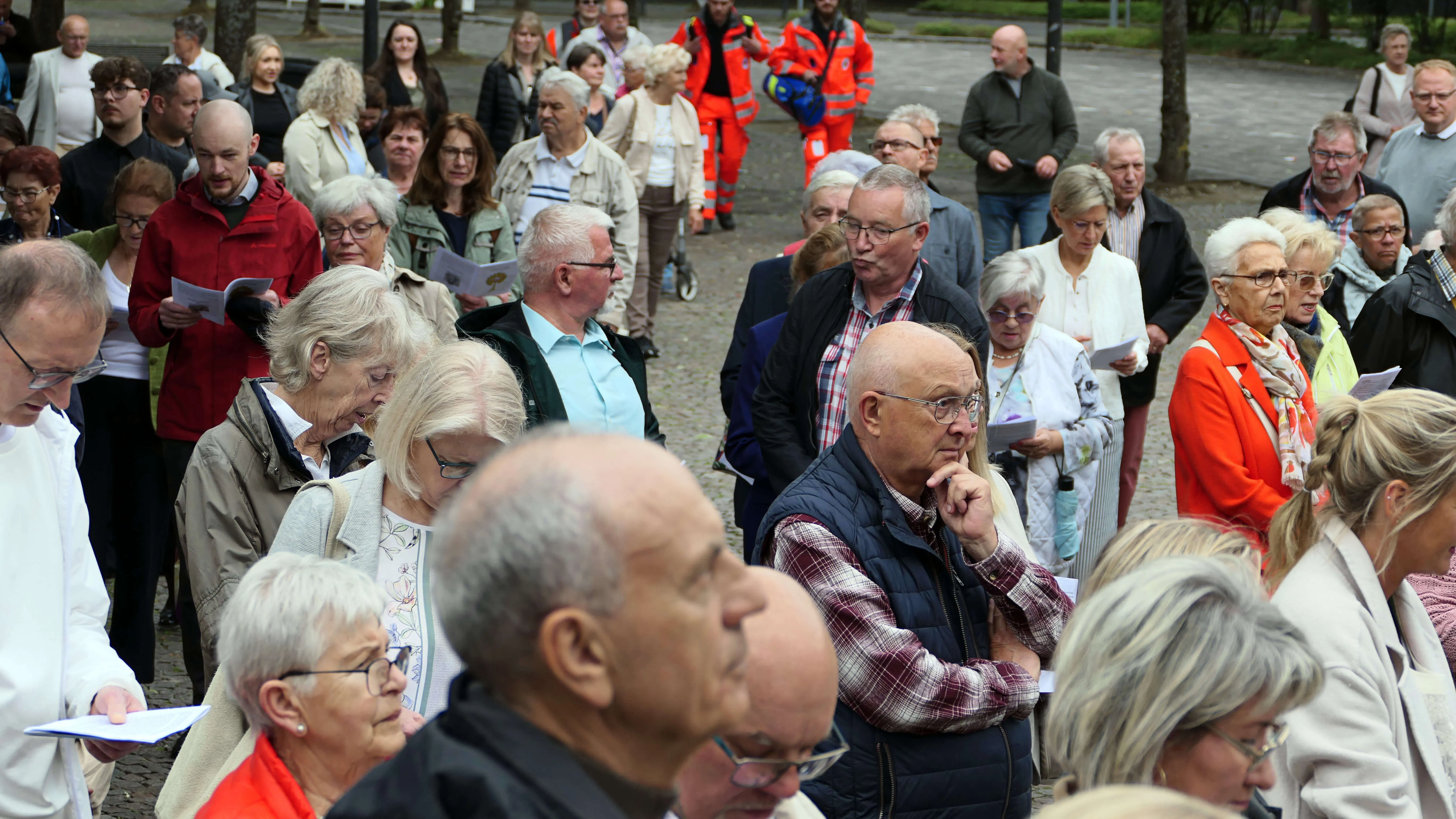 Eine große Menschenmenge steht und sitzt auf einem Platz vor einer Kirche, viele halten Blätter oder andere kleine Gegenstände in den Händen. Im Hintergrund sind weitere Personen und Bäume zu sehen.