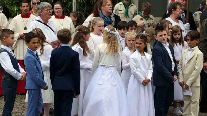 Kinder in weißen Gewändern und Anzugträger stehen während einer Prozession im Freien. Im Hintergrund sind weitere Menschen und ein Pfarrer in weißen Gewändern zu sehen.