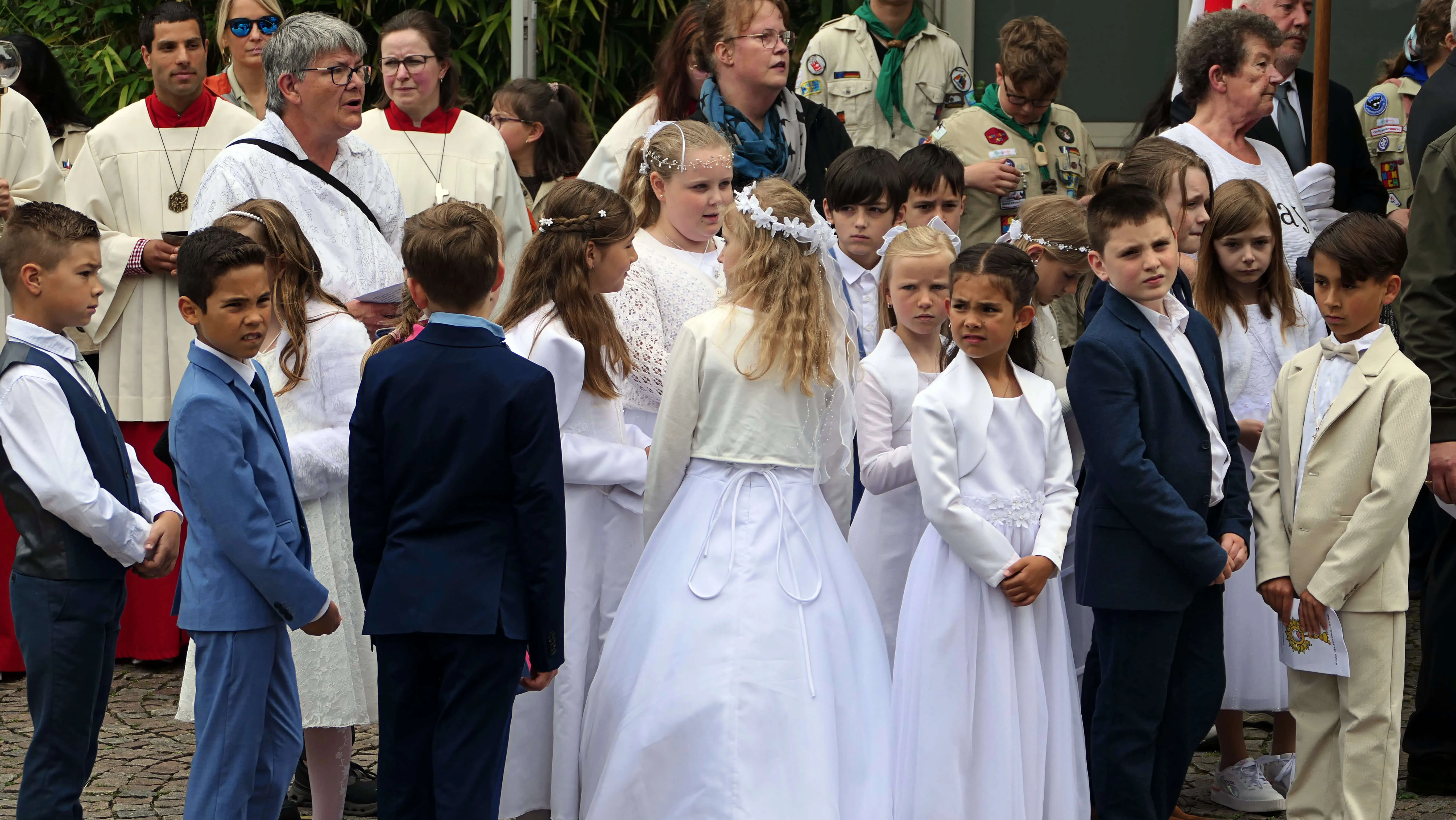 Kinder in weißen Gewändern und Anzugträger stehen während einer Prozession im Freien. Im Hintergrund sind weitere Menschen und ein Pfarrer in weißen Gewändern zu sehen.