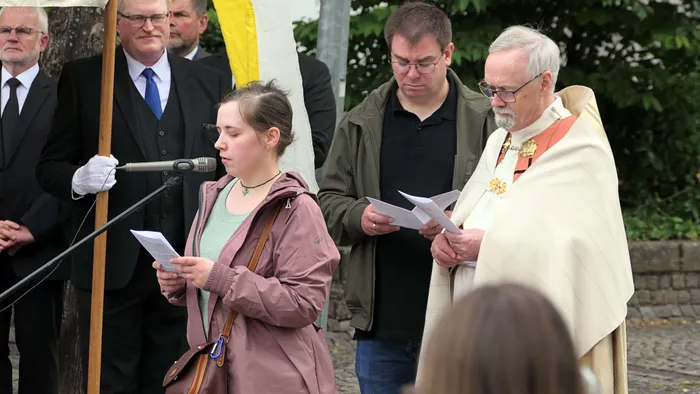 Eine junge Frau liest während einer Prozession aus einem Blatt Papier vor, während ein Priester in liturgischer Kleidung daneben steht und ebenfalls ein Blatt Papier hält.