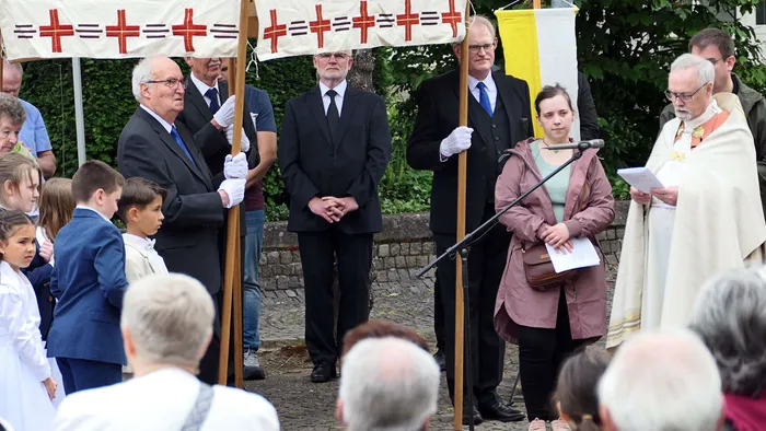 Eine Prozession mit Fahnen und Gläubigen vor einer Kirche. Ein Priester hält eine Ansprache, während andere Teilnehmer Fahnen tragen und zuhören.
