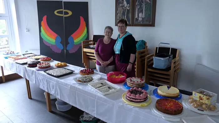 Zwei Frauen stehen hinter einem reich gedeckten Tisch mit Kuchen und Torten in einem Gemeindehaus. Im Hintergrund ein farbiges Wandbild mit religiöser Symbolik.
