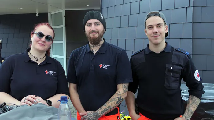 Drei ehrenamtliche Helfer des Deutschen Roten Kreuzes stehen vor einer grauen Wand. Sie tragen schwarze Poloshirts mit dem DRK-Logo und wirken aufmerksam.
