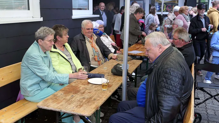 Mehrere ältere Menschen sitzen an Biertischbänken und unterhalten sich bei Getränken im Freien, während im Hintergrund eine Menschenmenge zu sehen ist.