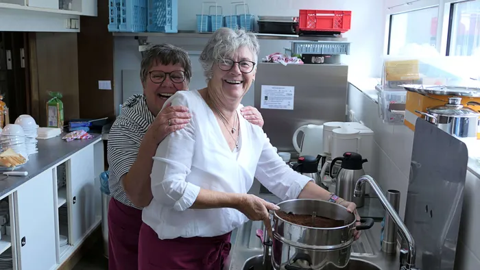 Zwei ältere Frauen lächeln in einer Küche und bereiten gemeinsam Speisen zu, während im Hintergrund ein Pfarrfest vorbereitet wird.