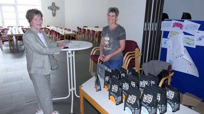 Zwei Frauen stehen an einem Tisch mit Kaffeetassen und mehreren Packungen Kaffee mit der Aufschrift „Tatico Fair Coffee“. Im Hintergrund sind Tische mit weißen Tischdecken und Stühle zu sehen.