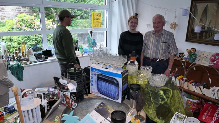 Drei Personen stehen an einem Tisch mit verschiedenen Gegenständen auf einem Flohmarkt oder Pfarrfest vor einem Fenster.
