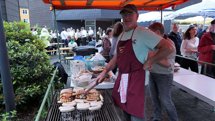 Mann grillt Würstchen auf einem Holzkohlegrill vor einer Menschenmenge bei einem Pfarrfest im Freien.