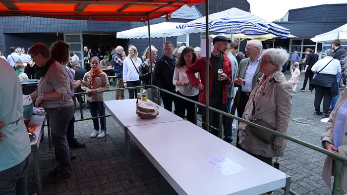 Menschen stehen an Tischen mit Getränken und Speisen vor einem Gebäude mit Markisen. Im Hintergrund sind weitere Menschen und ein Grill zu sehen.