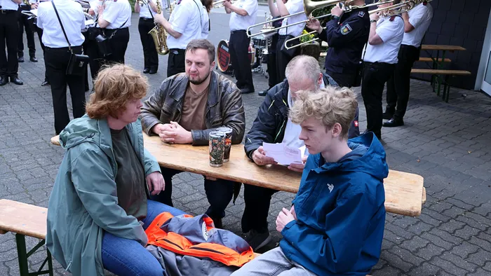 Vier Personen sitzen an einem Holztisch im Freien, während im Hintergrund eine Blaskapelle spielt. Sie unterhalten sich und haben Getränke vor sich.