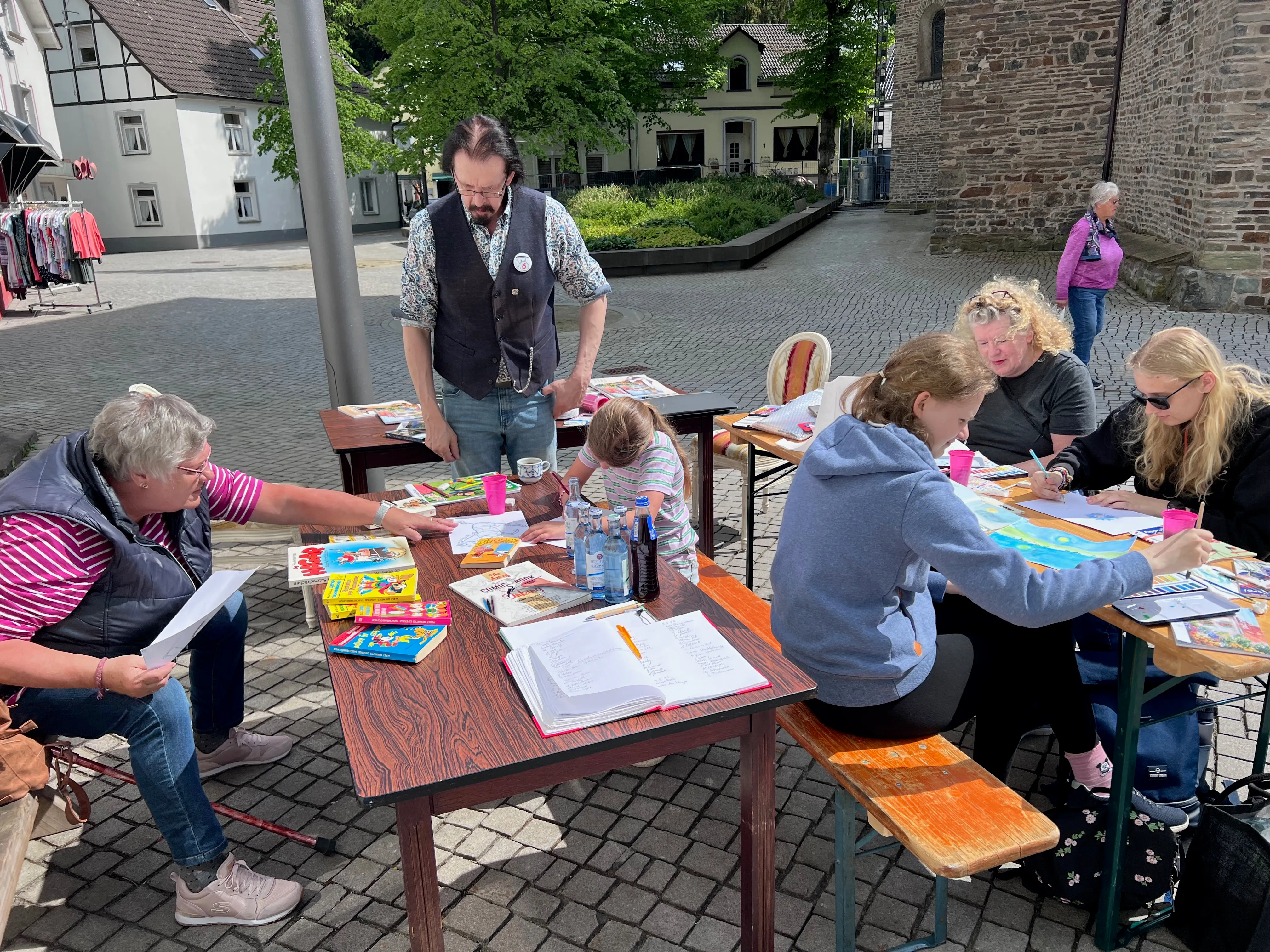 Menschen sitzen an Tischen und malen oder betrachten Comicbücher auf einem Frühlingsmarkt vor einem historischen Gebäude. Ein Mann mit Bart steht inmitten der Gruppe und hält einen Becher.