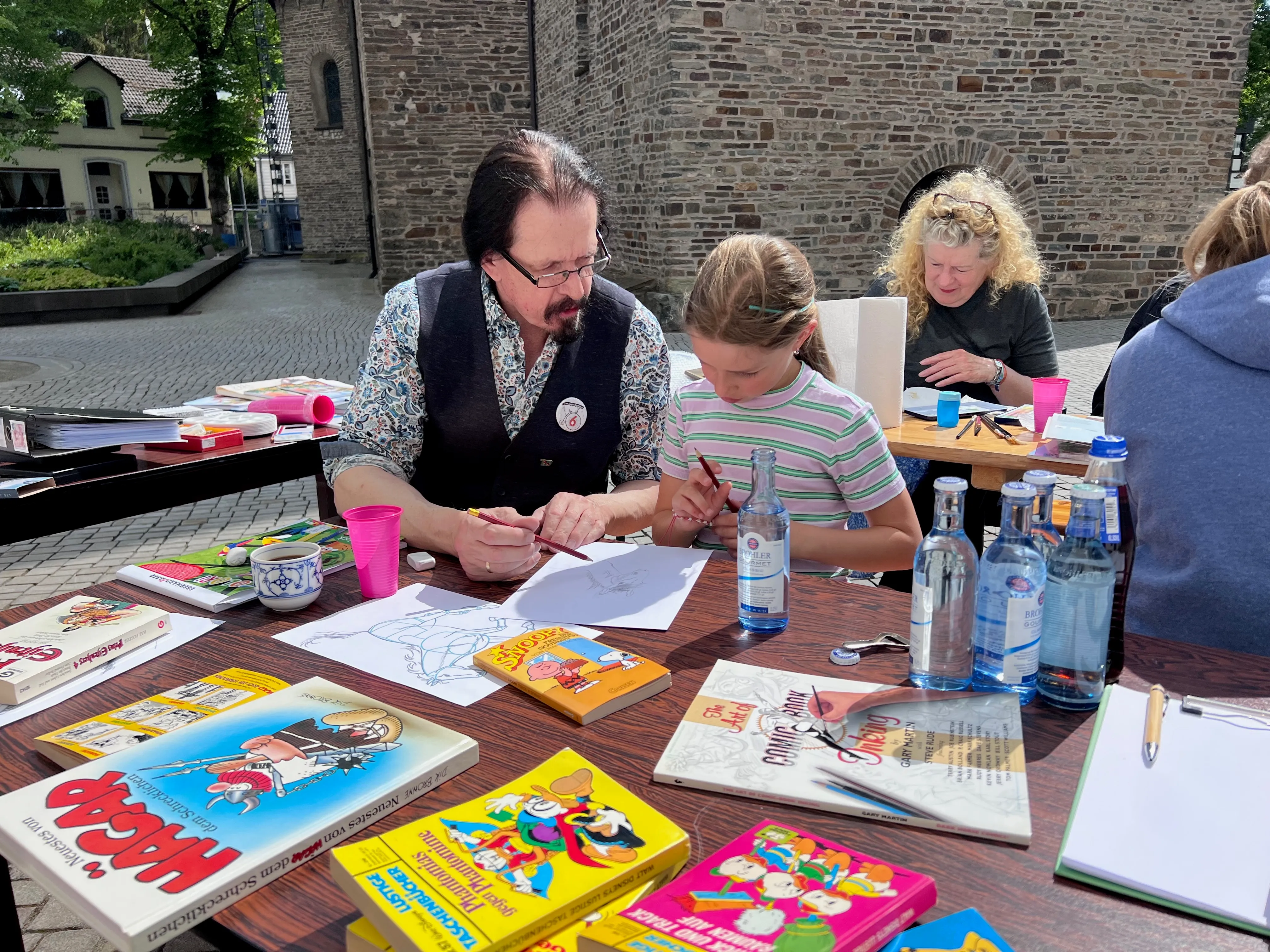 Comiczeichner Dietmar Krüger unterstützt ein Mädchen beim Malen am Tisch mit Comicbüchern und Malutensilien vor dem Heimathaus in Plettenberg.